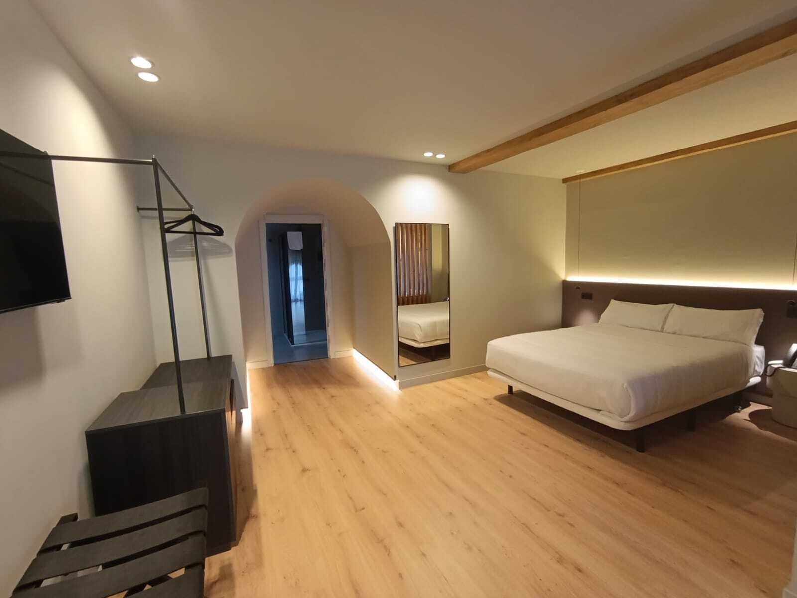 A modern hotel room with a large bed, a TV mounted on the wall, a wooden floor, and a mirror reflecting the room.