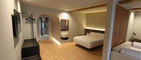 A modern hotel room with a wooden floor, a bed, a seating area with a table and chairs, a shower area, and a mirror.