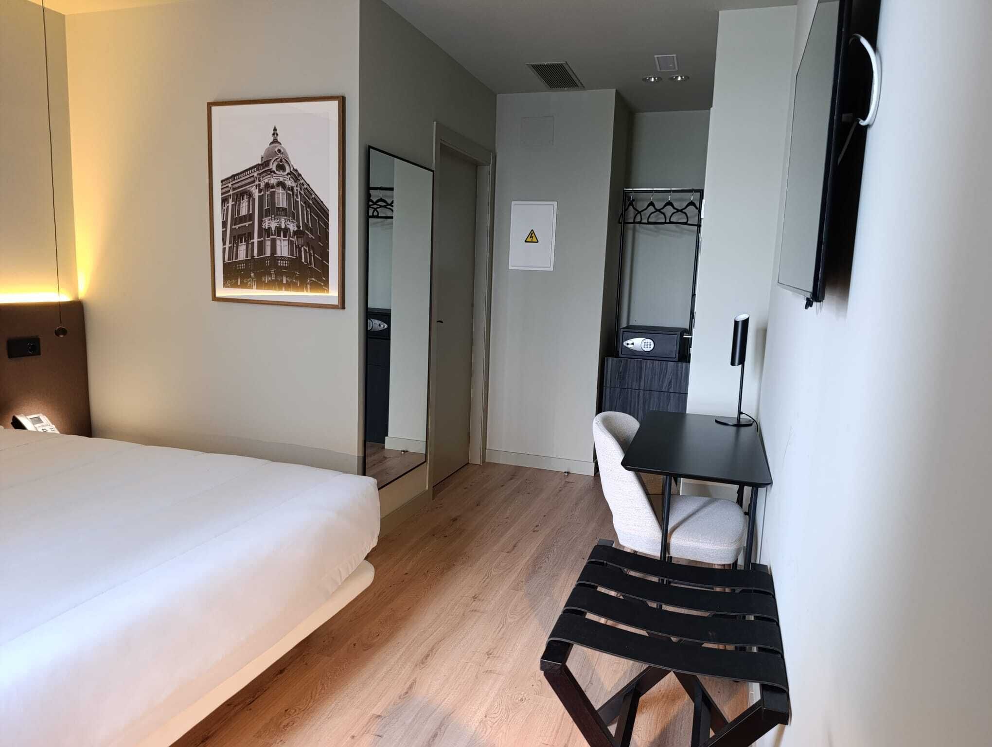 Habitación de hotel con cama, escritorio, silla, una mesita, espejo y un cuadro en la pared.