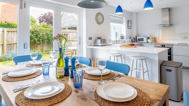Une cuisine moderne avec des meubles blancs, un îlot central et un coin repas agrémenté d’une table en bois pour quatre personnes.