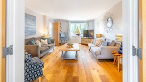 Un salon spacieux avec un canapé, une table basse et un téléviseur. Il y a une fenêtre avec des rideaux et une décoration sur le thème d’un voilier.