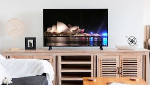 A flat-screen TV on a wooden TV stand displaying the Sydney Opera House at night. - Zen Coastline Pool - Balcony Dining - Seaview (Darwin)
