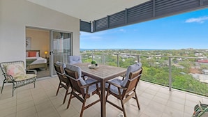 A balcony with a wooden table and chairs, a bed with a view, and a glass door leading to an interior room. - Zen Coastline Pool - Balcony Dining - Seaview (Darwin)