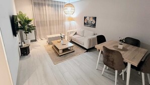 A modern living room with a sofa, coffee table, dining table, and chairs. There is a TV mounted on the wall, a potted plant, and a painting on the wall. - Charmant Appart Punique au Cœur de Carthage (Carthage)