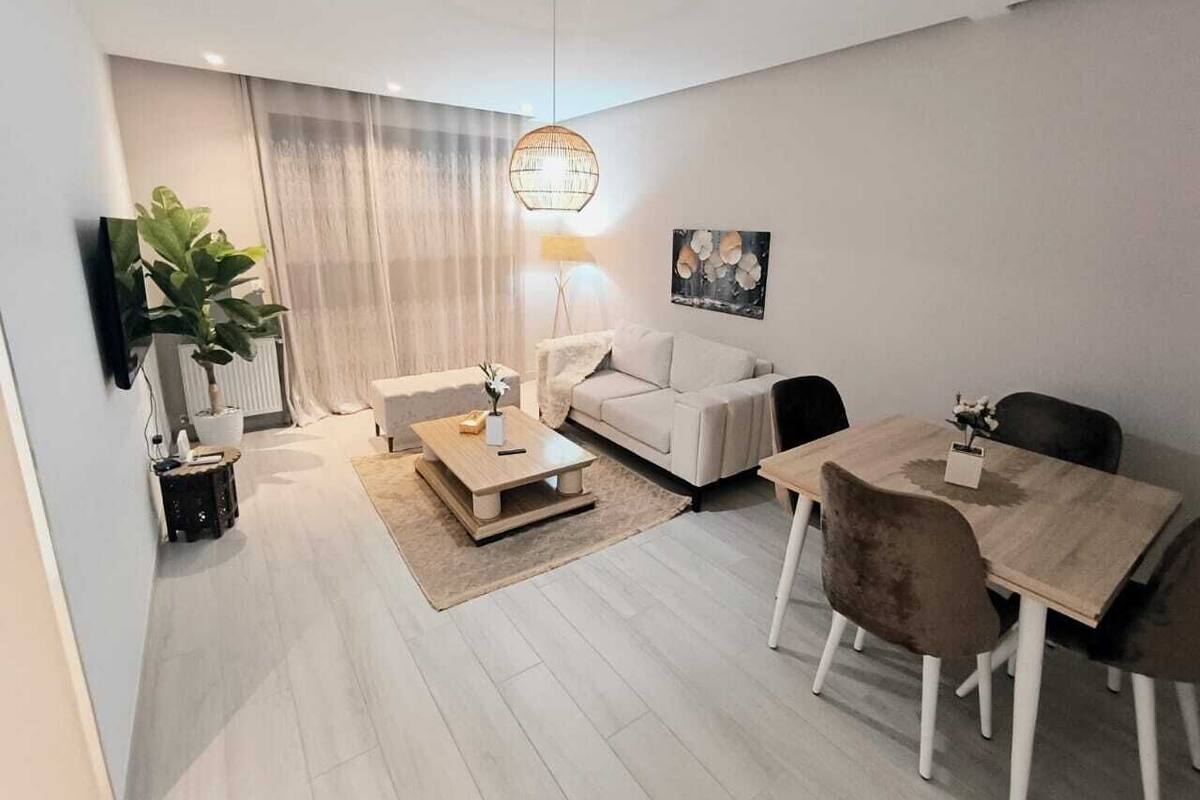 Ett modernt vardagsrum med en soffa, ett soffbord, ett matbord och stolar. En tv är monterad på väggen, och det finns en krukväxt och en tavla på väggen.