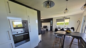 A modern kitchen with built-in appliances, a dining area with a large table and bar stools, and a view of the outdoors through the windows.