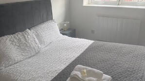 Una cama bien hecha con cabecera gris y ropa de cama blanca, una mesita de noche con lámpara, y una ventana con vista a edificios.