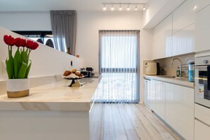 Una cocina moderna con gabinetes blancos, encimera de mármol y un amplio ventanal con cortinas ligeras.