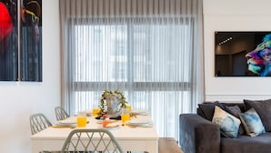 Un salon moderne avec un canapé foncé, une table à manger avec des chaises et une grande fenêtre équipée de stores.