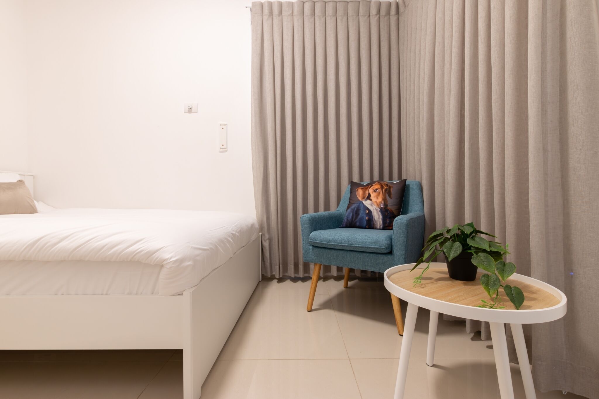 Una habitación de hotel con una cama, un sillón azul, una mesita redonda con una planta y una ventana con cortinas.