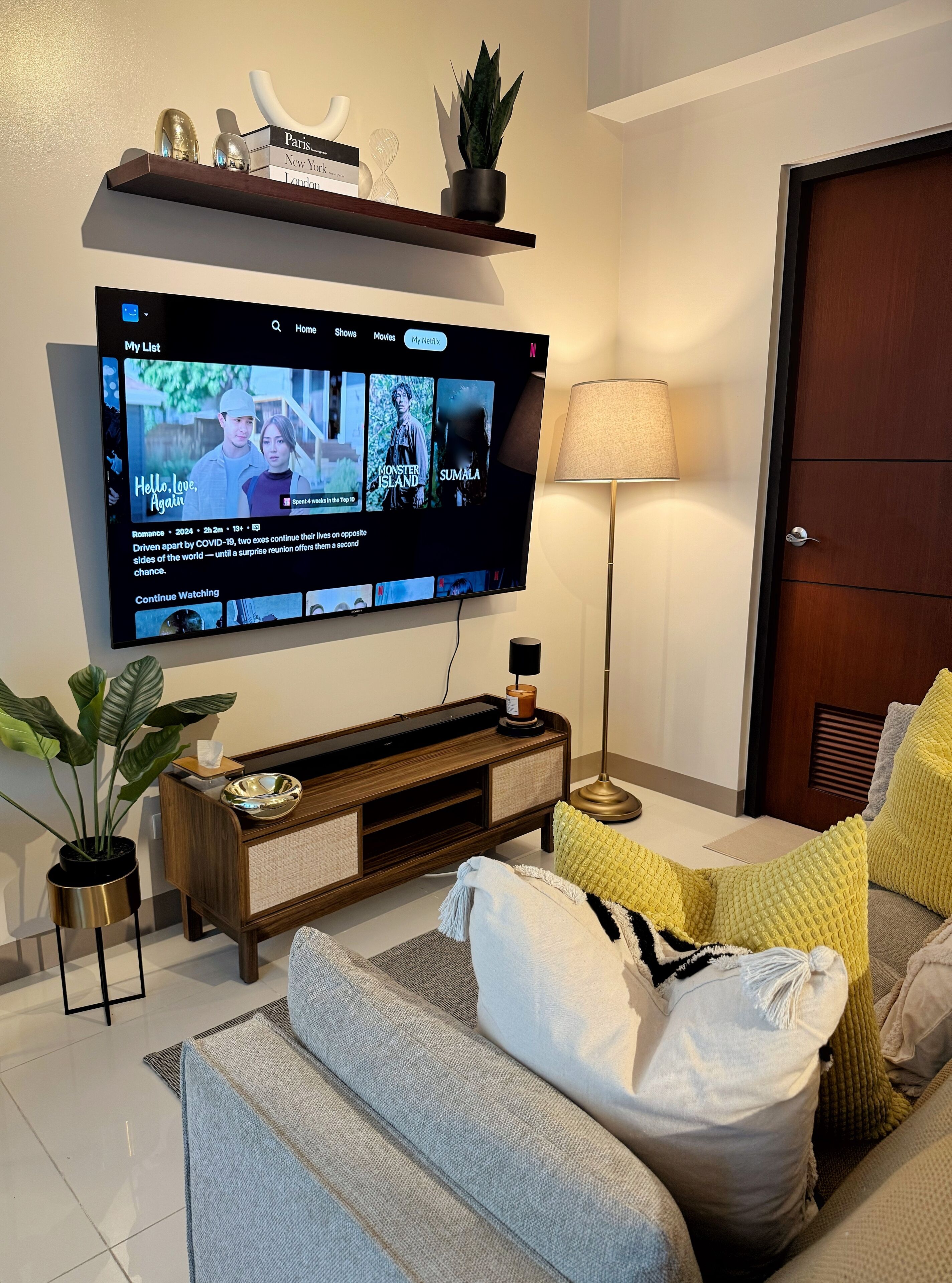 Una sala de estar moderna con un televisor de pantalla plana montado en la pared, una mesa consola de madera con objetos decorativos, una lámpara de pie y un sofá con cojines.