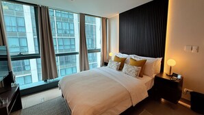 Et moderne hotelværelse med en stor seng, et natbord, en lampe og udsigt over byen gennem store vinduer.