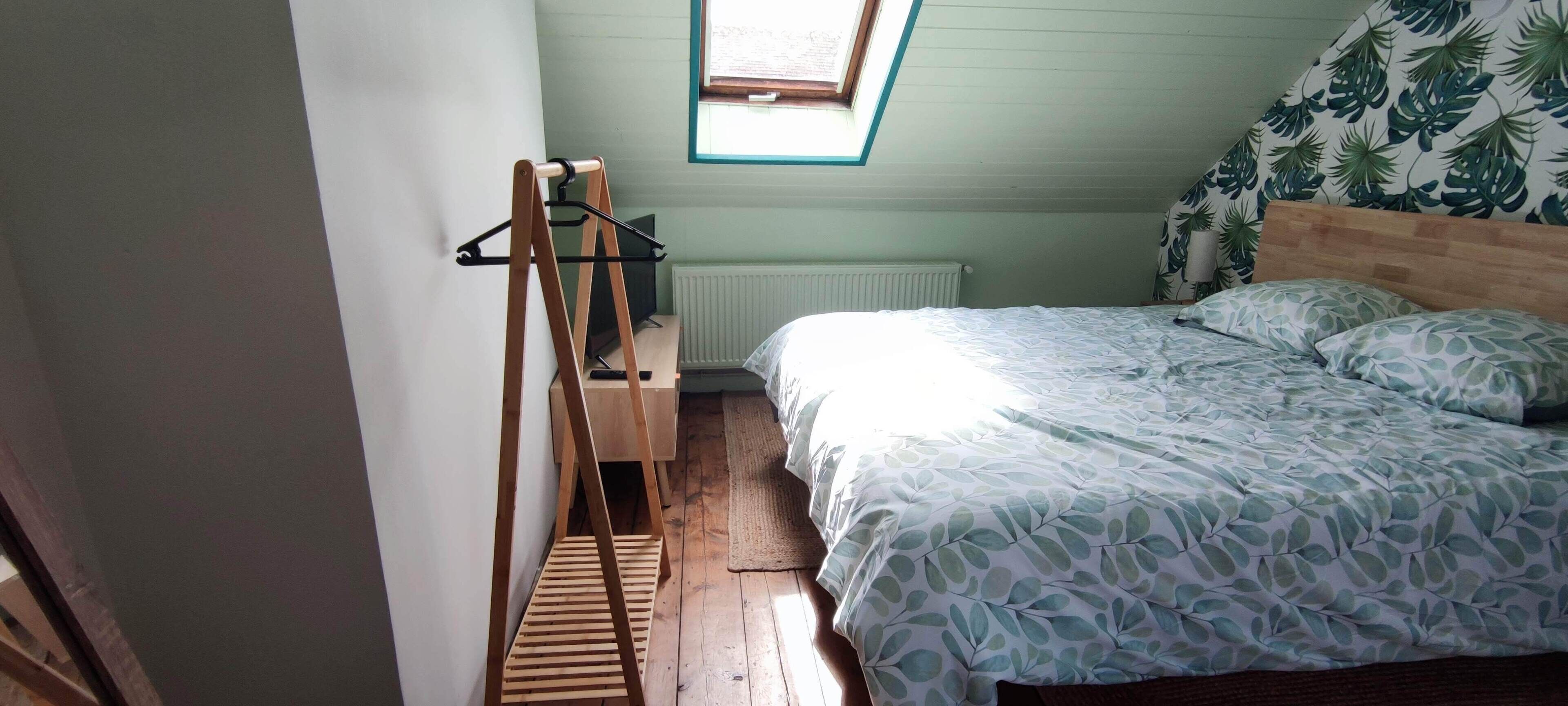 Ein Schlafzimmer mit schrägen Wänden, einem Bettgestell aus Holz, einem Heizkörper und einem Fenster mit Blick auf den Himmel.