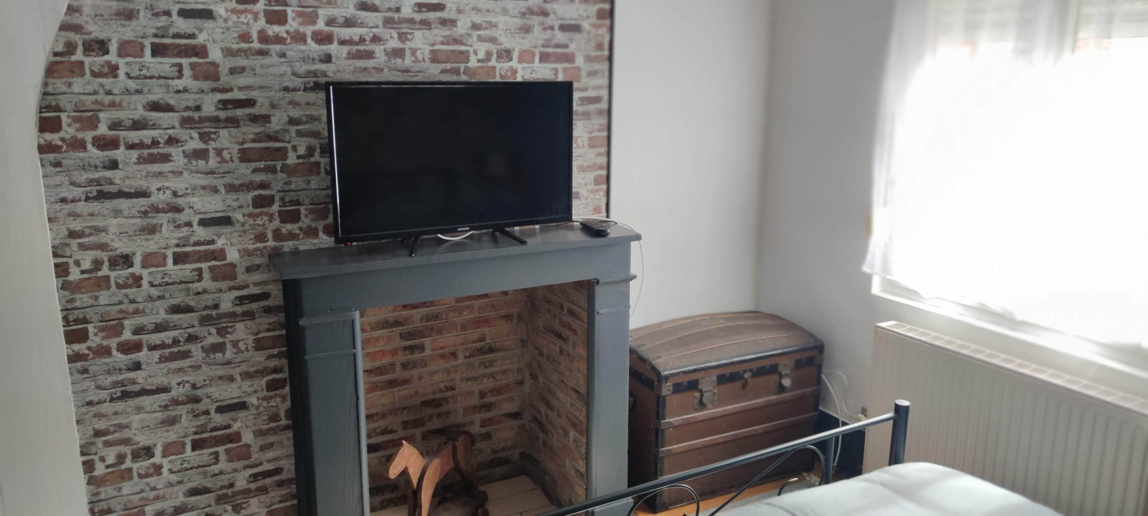Una habitación con chimenea de ladrillo, un televisor sobre un pedestal, un cofre de madera y una ventana con cortinas.