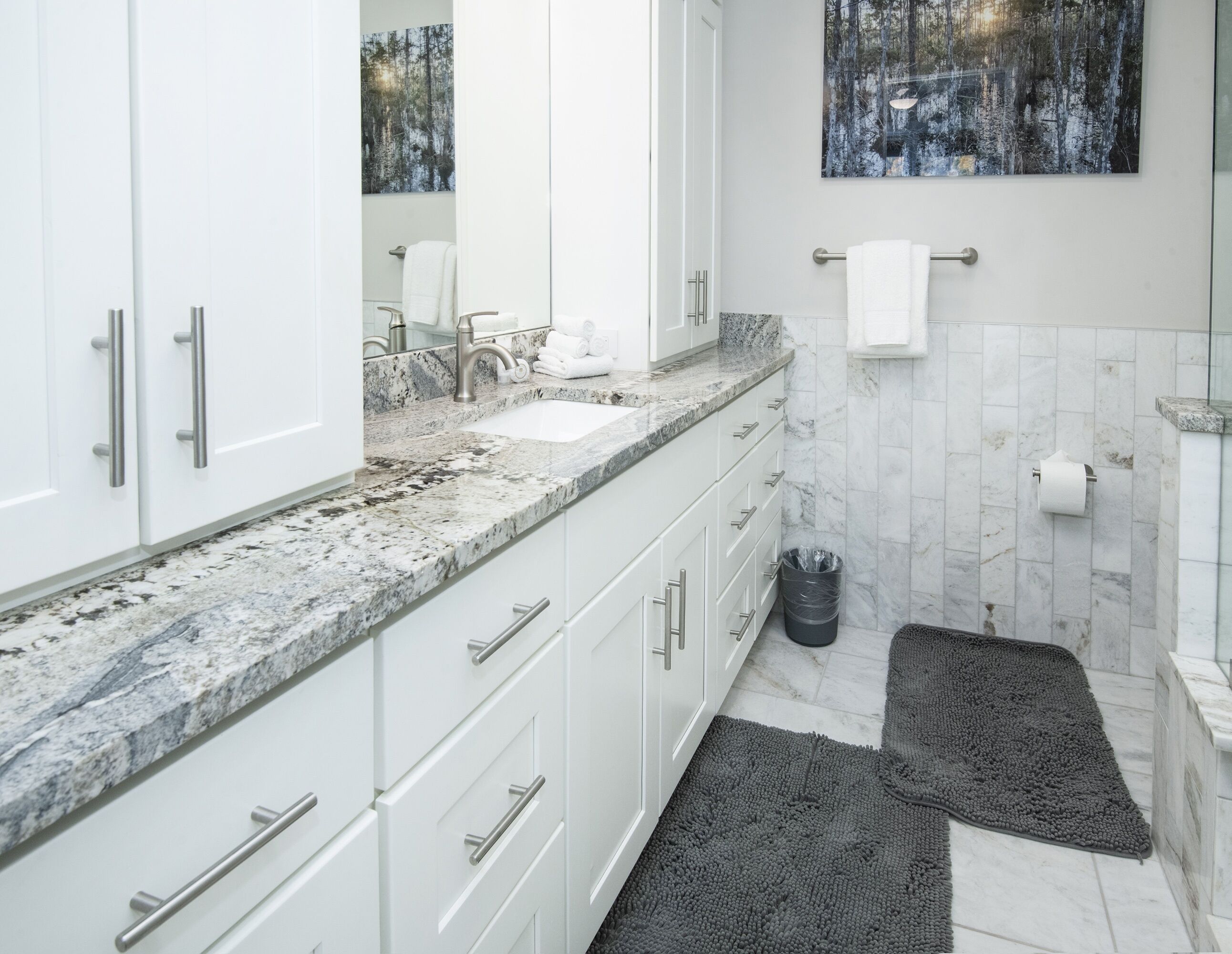 Un baño moderno con muebles blancos, encimera de granito, lavamanos, espejo y toallero.