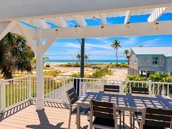 A covered outdoor deck with a view of the beach, a table with chairs, and a white railing. - 5550 Palmetto Paradise: 3br W/ Gulf Views, Heated Pool 3 Bedroom Home (Fort Myers Beach)