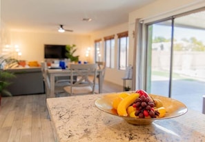 A kitchen with a fruit bowl on the counter, a dining area with chairs, and a television in the background. - Casa Alessia 4br Home & Heated Pool 4 Bedroom Home (Casa Grande)