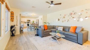 Ein modernes Wohnzimmer mit einem grauen Sofa, orangefarbenen Kissen und einem Couchtisch. Es gibt einen Deckenventilator, eine Wand mit runden Verzierungen und eine Küche im Hintergrund.