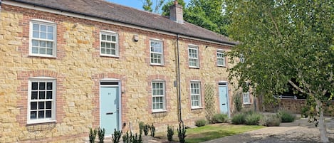 A stone building with multiple windows and doors, a gravel driveway, and a small garden area with shrubs.