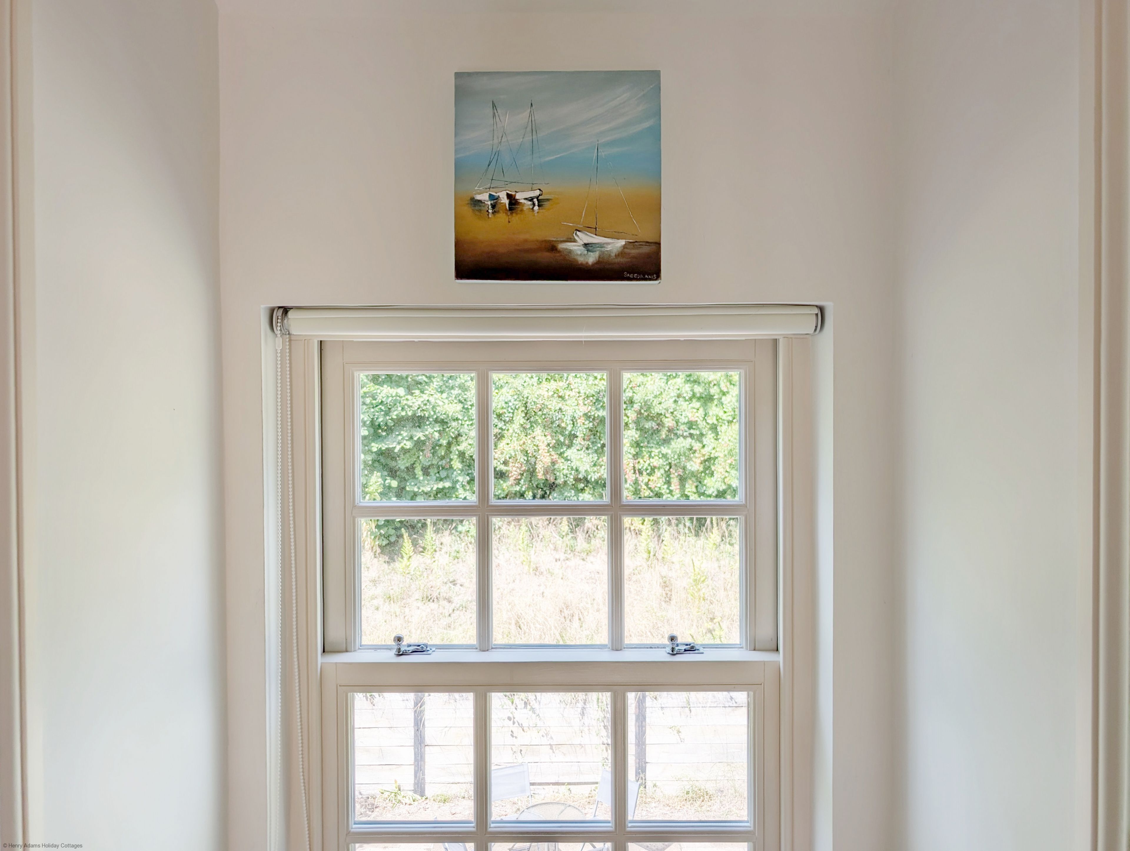 Una habitación con un ventanal grande, un cuadro de veleros arriba y vistas al exterior con follaje.