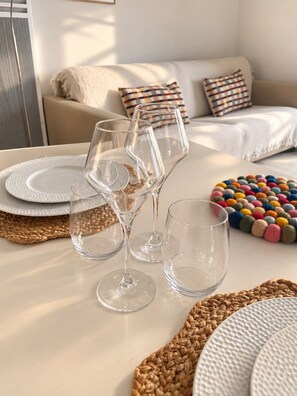 A dining table with clear glassware and woven placemats, a colorful pom-pom ball arrangement, and a neutral-toned sofa in the background. -  2 ROOM 3* FLAT WITH A/C & POOL  
BORMES/LE LAVANDOU (Bormes-les-Mimosas)