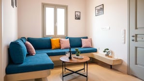 Een woonkamer met een blauwe bank, een salontafel en een lange bank. Er zijn twee ramen, een deur en lichtschakelaars op de muur.