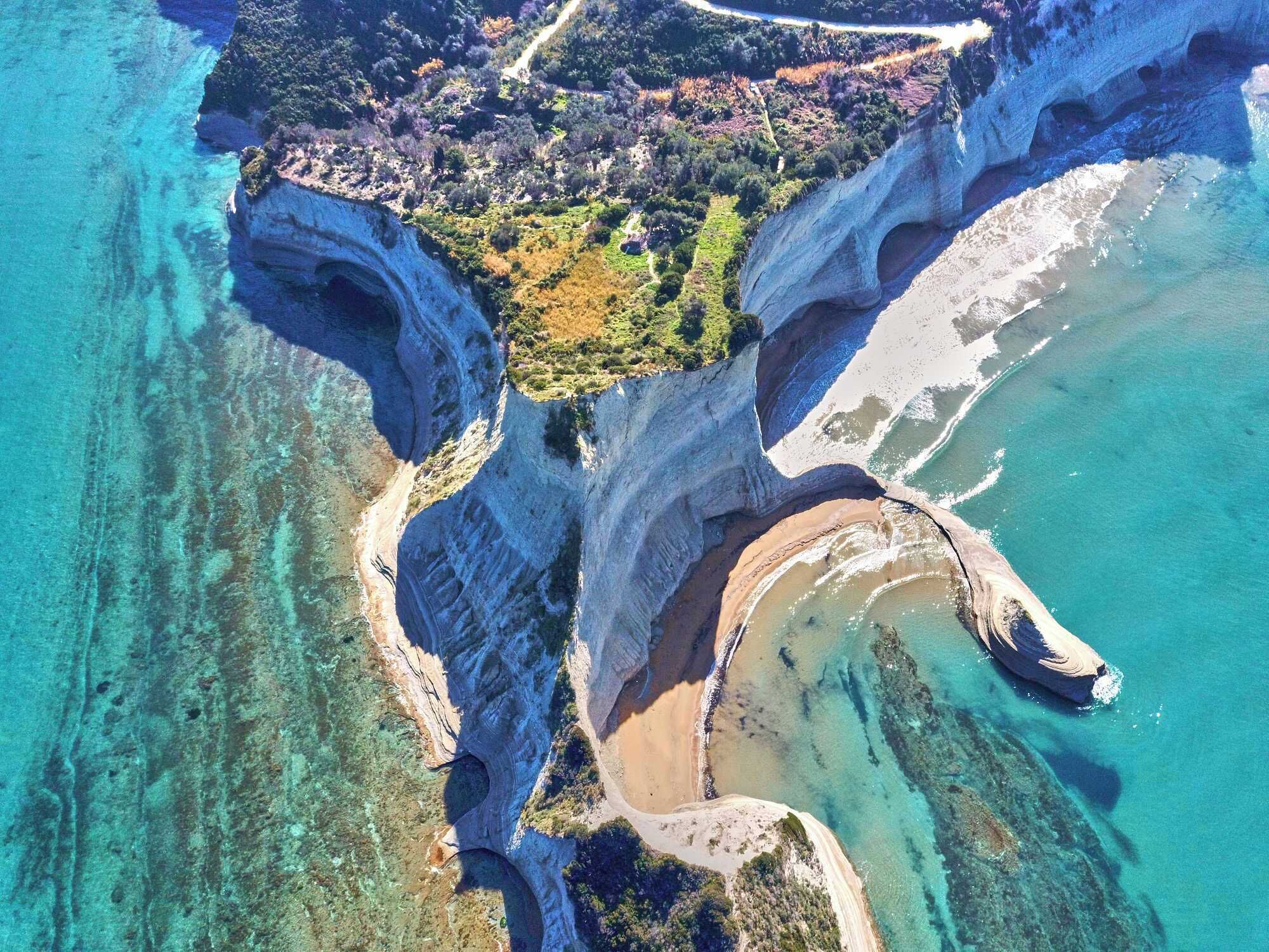 An aerial view of a coastal area with cliffs, clear waters, and a small patch of vegetation.