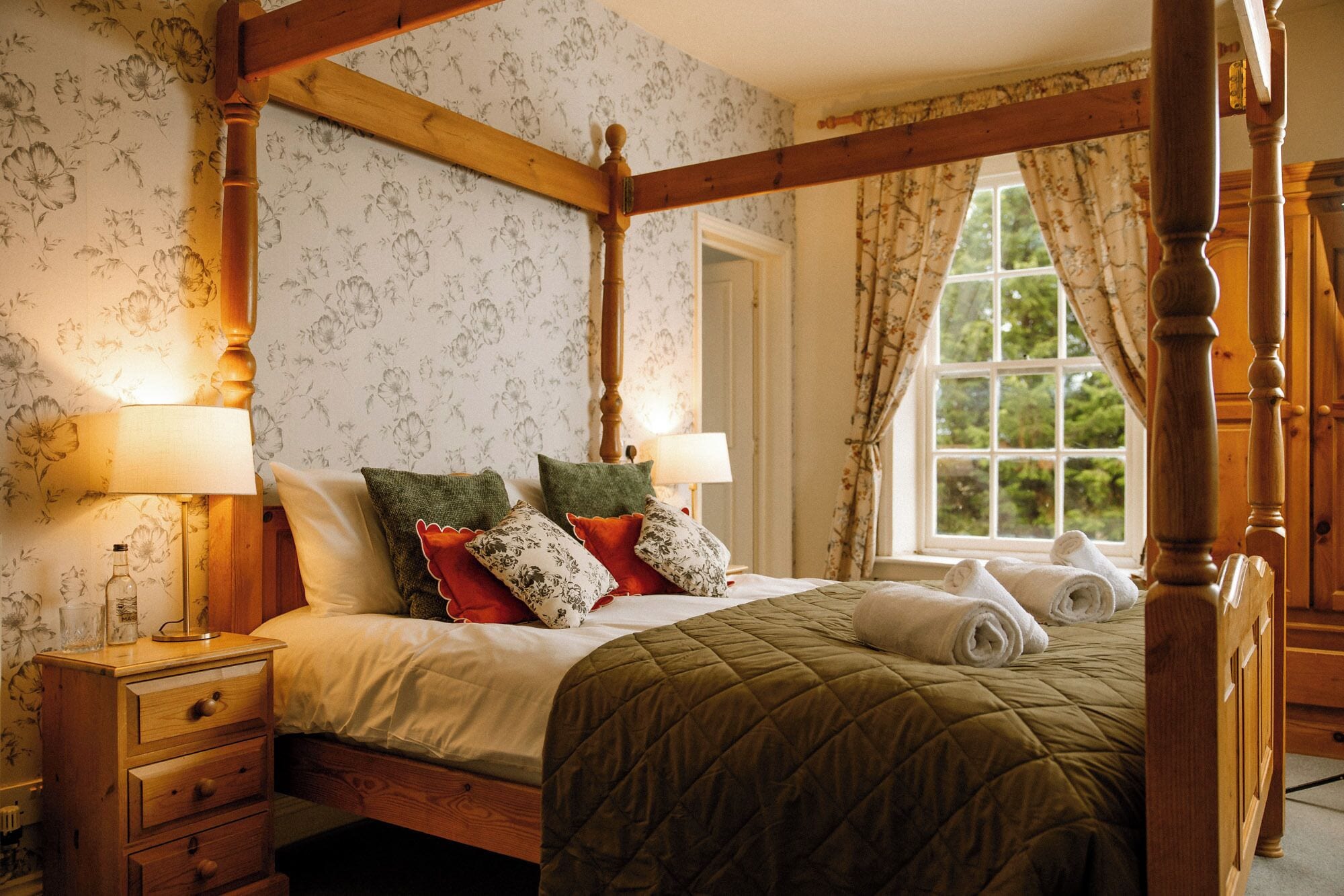 A four-poster bed with a quilted bedspread, two bedside lamps, and floral wallpaper.