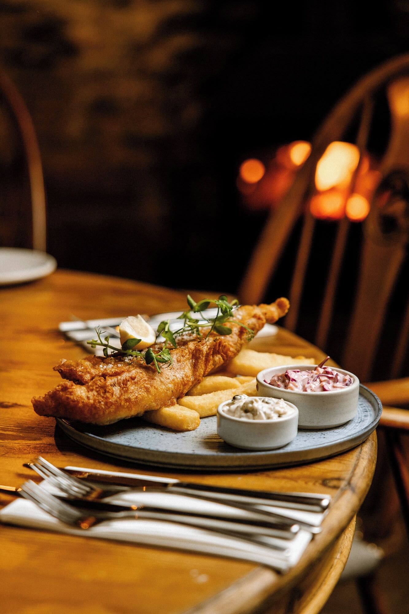A plate of fish and chips with lemon wedges and a side of coleslaw on a wooden table.