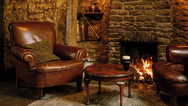 A cozy room with a stone fireplace, leather armchairs, and a small table with a drink. - The George Inn (Lower Brailes)