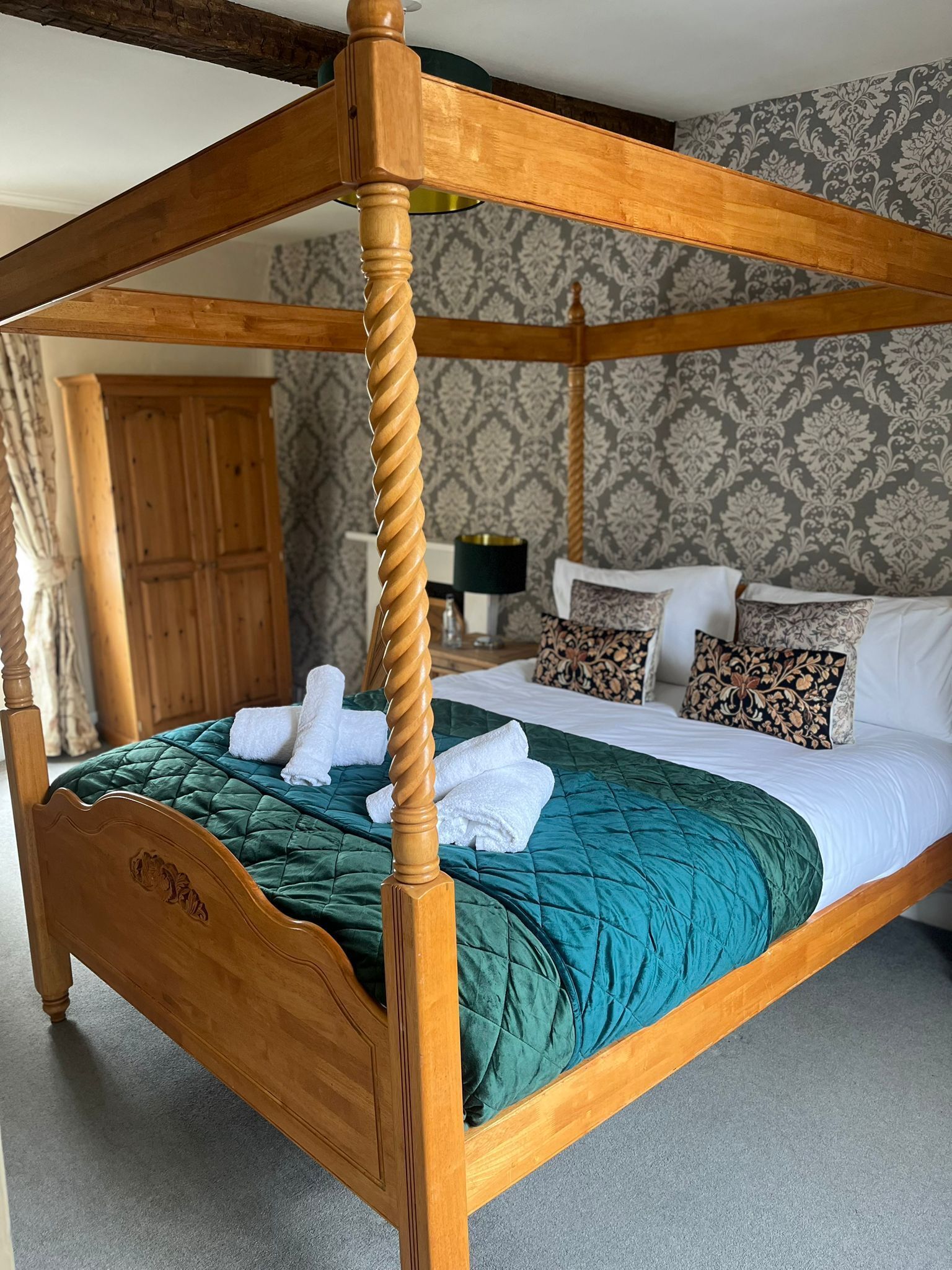 A four-poster bed with a green quilt and white towels, a wooden headboard, and patterned pillows.
