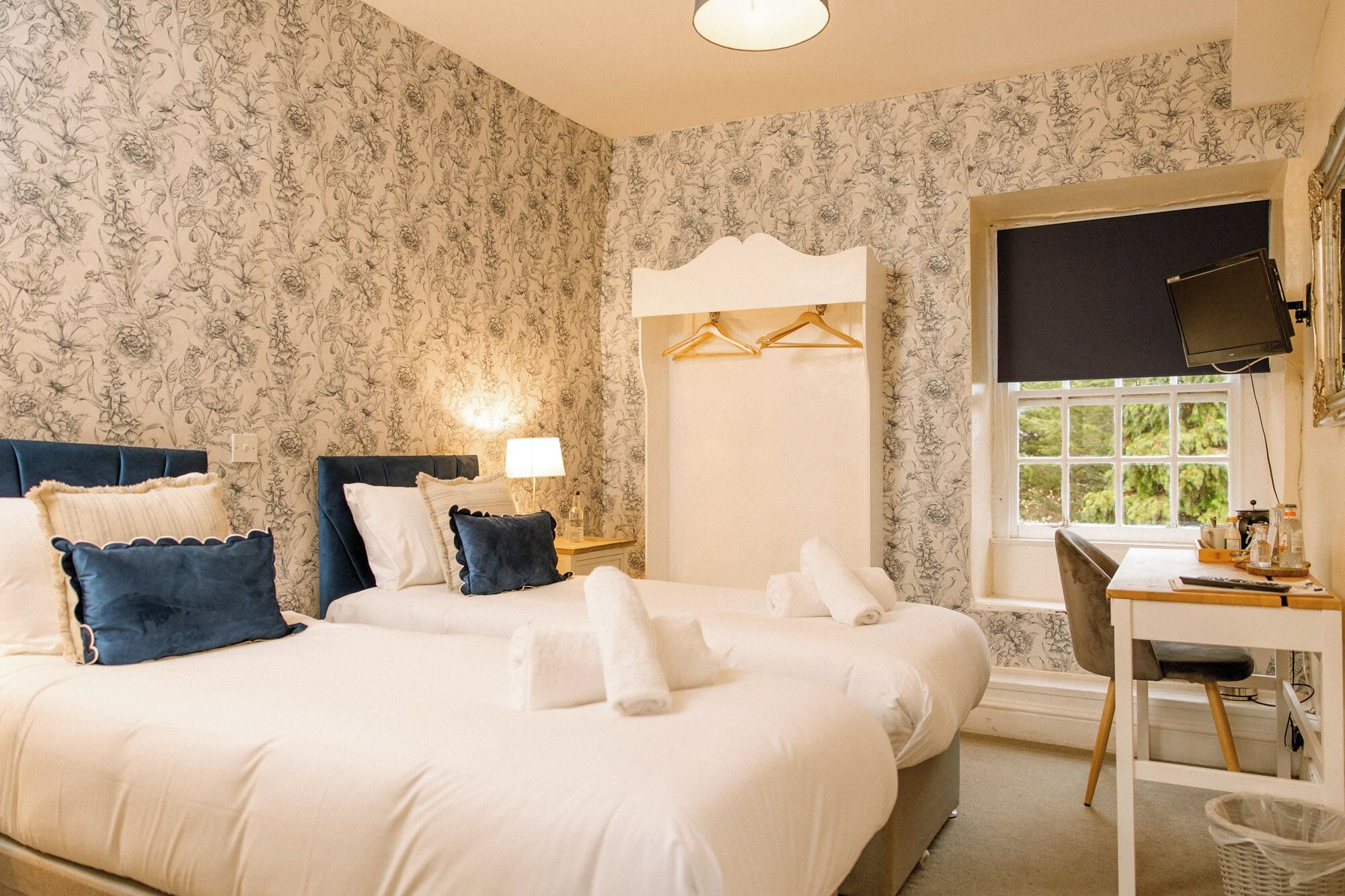 A hotel room with a large bed, a desk with a chair, a window with a view of greenery, and a wall with floral wallpaper.