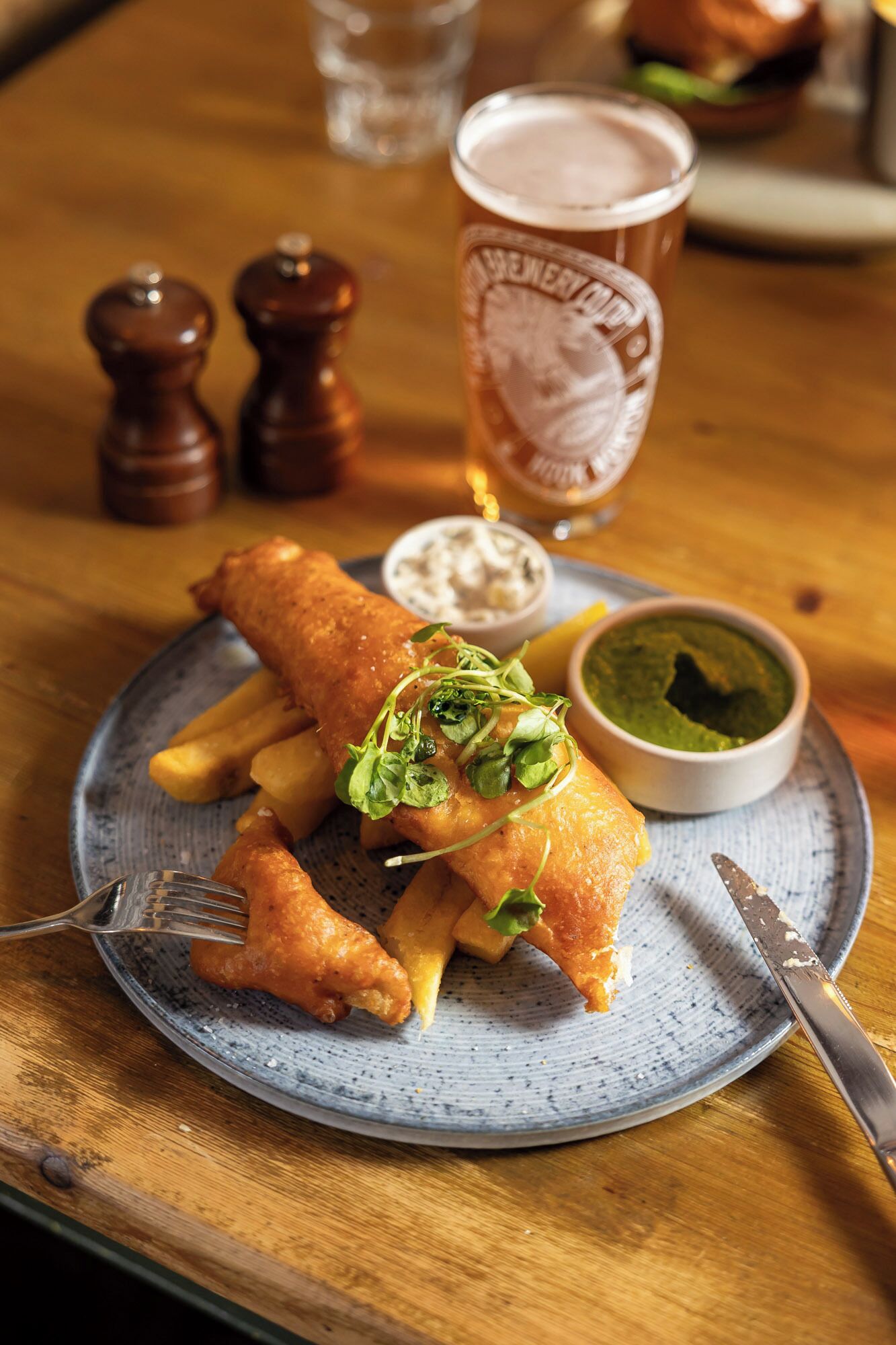 A plate of fish and chips with a side of tartar sauce and a green sauce, a glass of beer, and salt and pepper shakers on a wooden table.