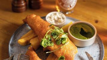 A plate of fish and chips with a side of tartar sauce and a green sauce, a glass of beer, and salt and pepper shakers on a wooden table.