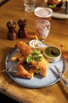 A plate of fish and chips with a side of tartar sauce and a green sauce, a glass of beer, and salt and pepper shakers on a wooden table.