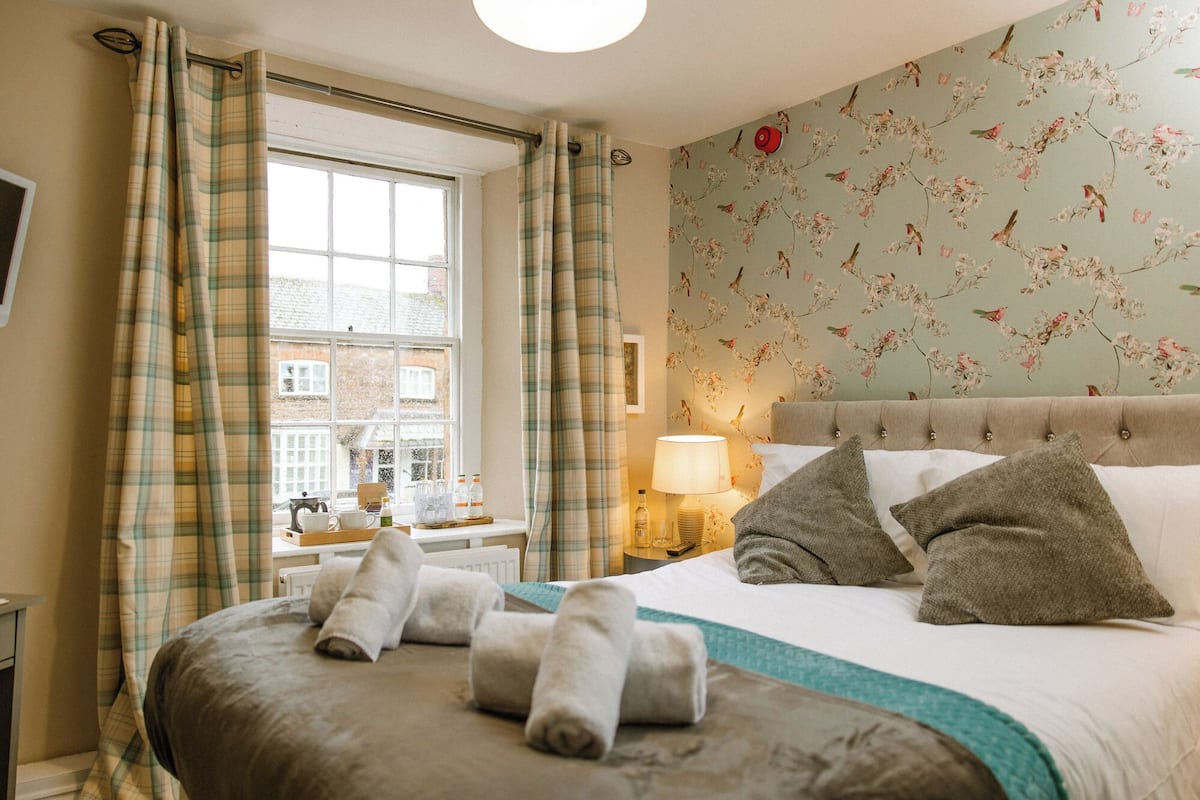 Une chambre à coucher avec un grand lit, une fenêtre avec des rideaux, un papier peint à motifs floraux et une table de chevet avec une lampe.