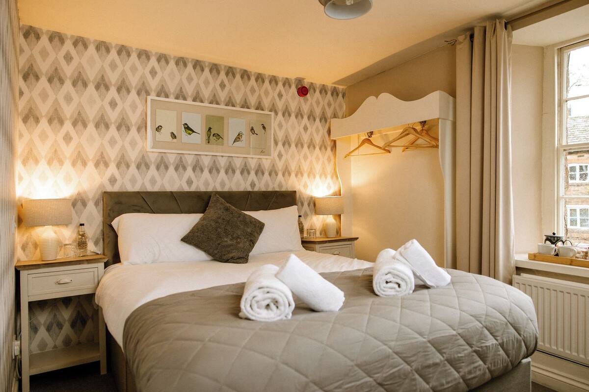 Une chambre d’hôtel avec un lit, deux tables de chevet, un tableau représentant des oiseaux accroché au mur, une fenêtre avec des rideaux et un radiateur.