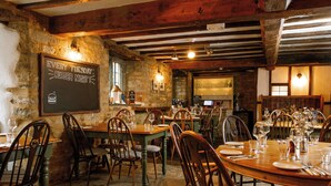 A restaurant with rustic stone walls, wooden beams, and a chalkboard sign advertising 'Every Tuesday Burger Night'. Tables are set with wine glasses and plates, and chairs are wooden with cushioned seats. - The George Inn (Lower Brailes)
