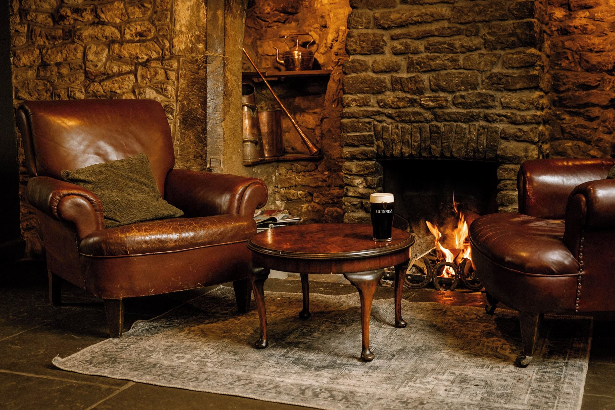 A cozy room with a stone fireplace, leather armchairs, a small table with a drink, and a fire burning.