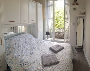 A bedroom with a bed, a nightstand, a lamp, a mirror, and a view of the outdoors.
