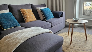 A modern living room with a grey sofa, patterned cushions, a small round table, and a view through a window. - Apartment facing the Royat thermal baths (Royat)