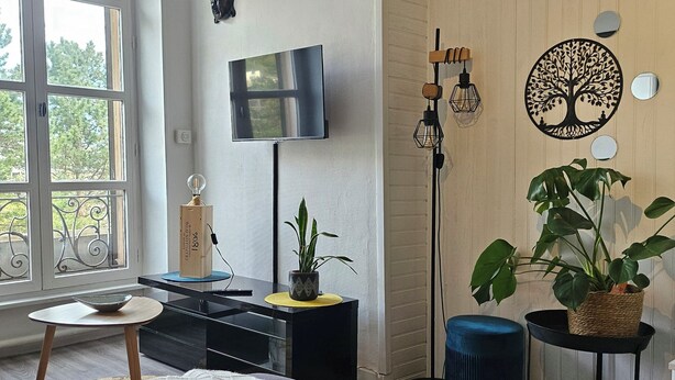 A modern living room with a flat-screen TV, a plant, a decorative tree wall art, a blue ottoman, and a wooden floor. - Apartment facing the Royat thermal baths (Royat)