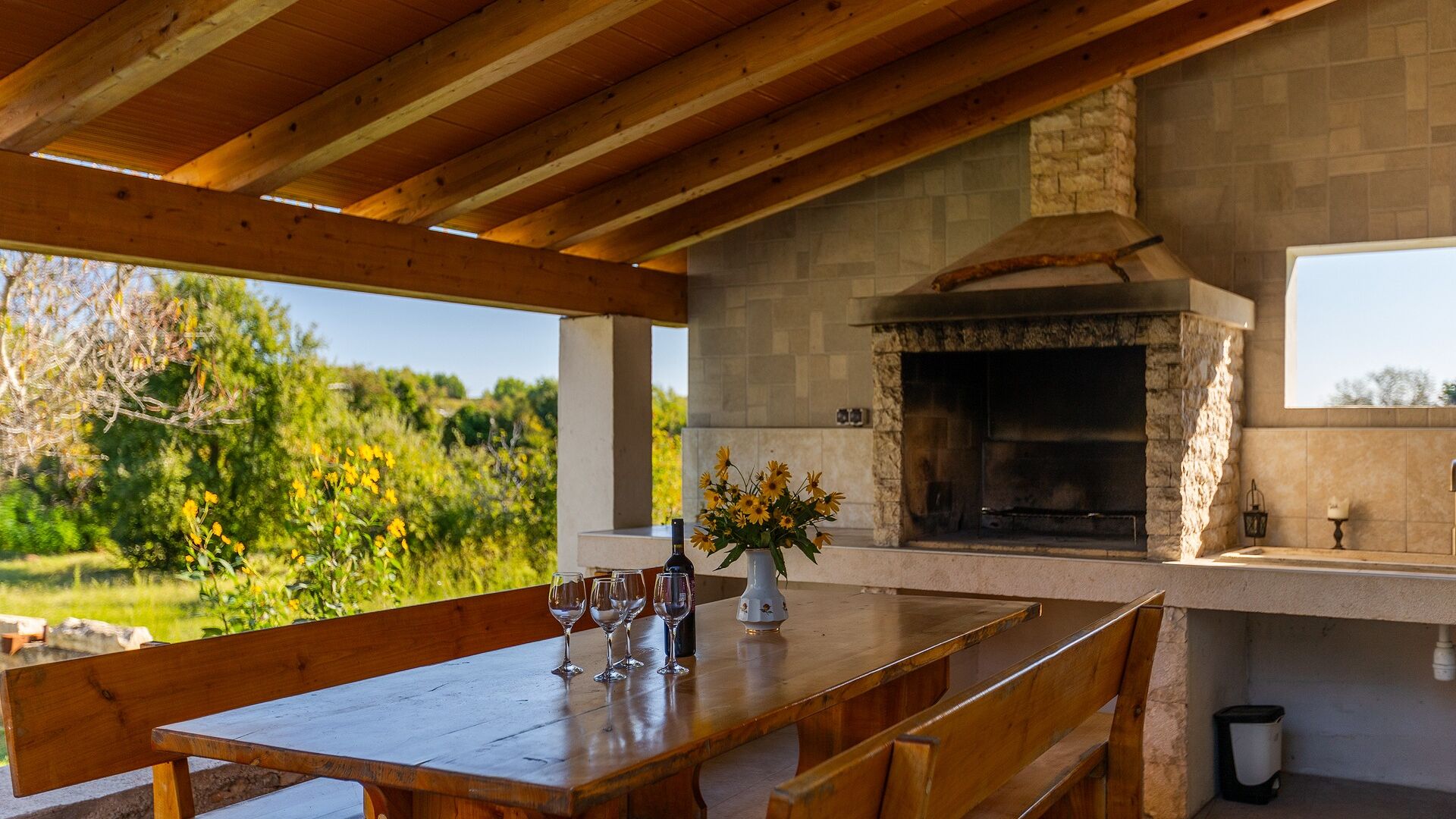 An outdoor dining area with a stone fireplace, wooden beams, and a view of greenery.