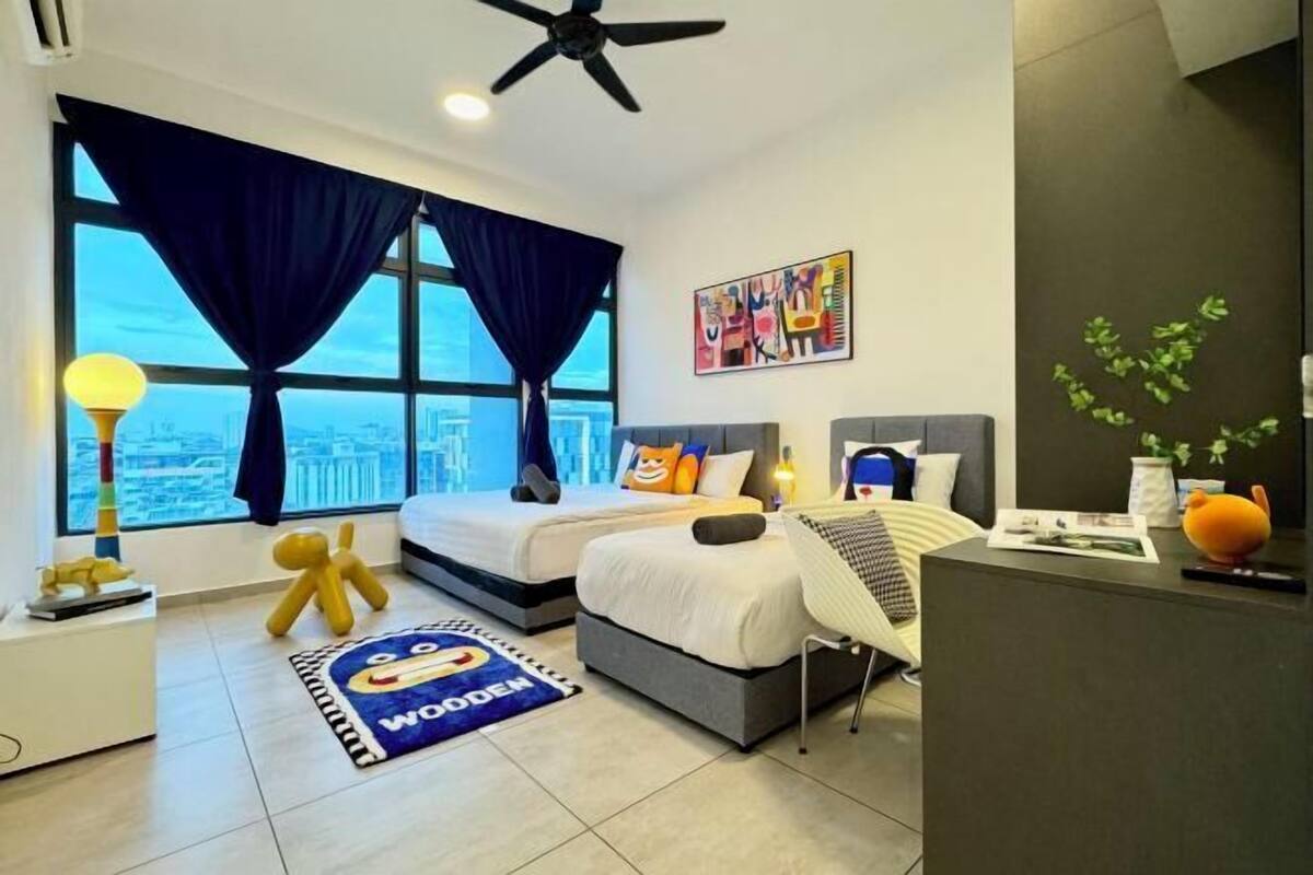 A hotel room with two beds, a desk, a ceiling fan, a large window with curtains, a city view, and a colorful artwork on the wall.