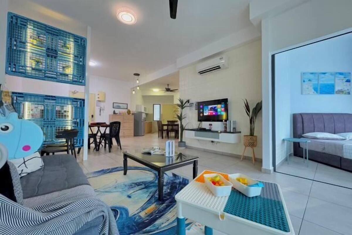A modern living room with a sofa, coffee table, and TV. There is a dining area with chairs and a table. The room has a ceiling fan and an air conditioning unit.
