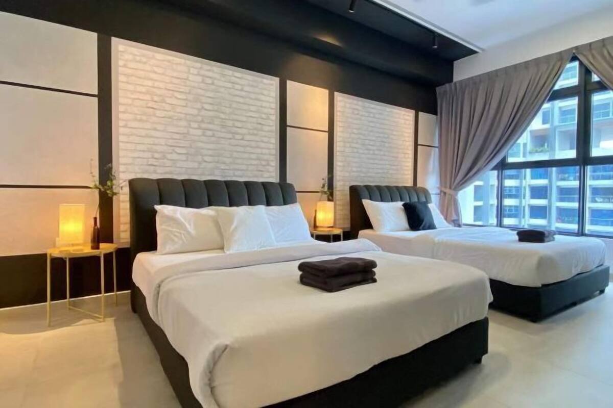 A hotel room with two beds, a large window with curtains, and a modern design with a brick wall accent.