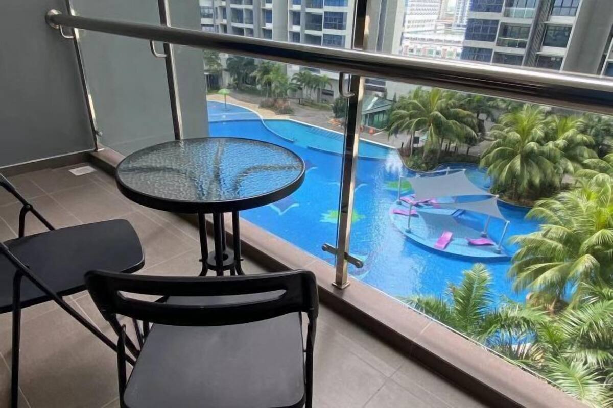 A balcony with a glass table and chairs, overlooking a swimming pool with palm trees and a view of surrounding buildings.
