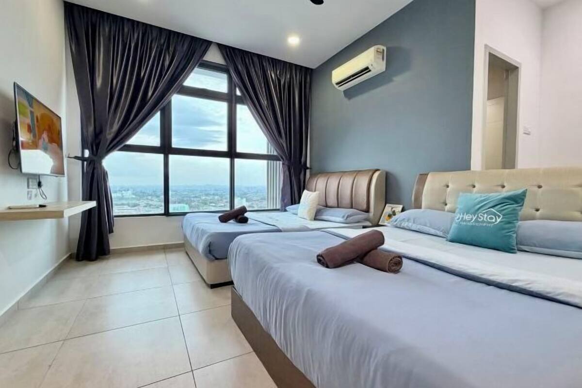 A hotel room with two beds, a large window with curtains, an air conditioning unit, and a view of the city.