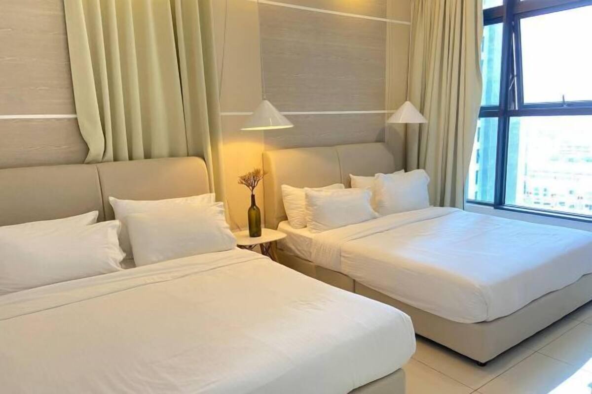 A hotel room with two beds, a small table with a vase, and a view of the city through large windows.
