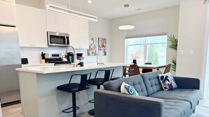 A modern kitchen with white cabinets, a central island with bar stools, a grey sofa with decorative pillows, and a dining area with a table and chairs. - 4BR Home Near Naperville! Parking + Pickleball (Warrenville)